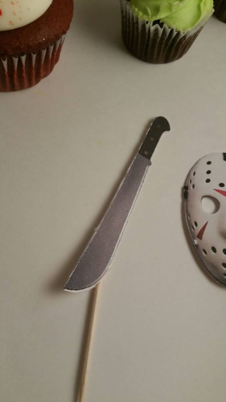 May include: A white cupcake with red frosting and a white cupcake with green frosting. A white paper machete and a white paper hockey mask are on a white surface.