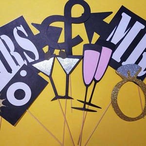 May include: A collection of black and white photo booth props on wooden sticks, including "MRS" and "MR" signs, champagne flutes, martini glasses, and a glittery engagement ring. The props are set against a yellow background.