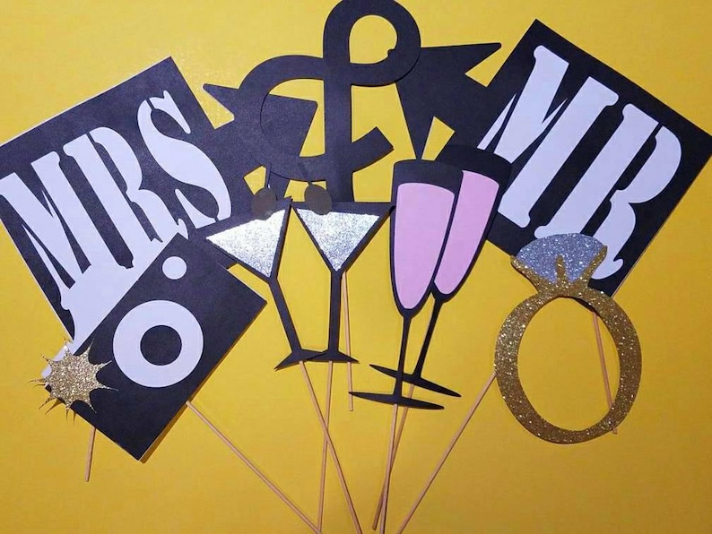 May include: A collection of black and white photo booth props on wooden sticks, including "MRS" and "MR" signs, champagne flutes, martini glasses, and a glittery engagement ring. The props are set against a yellow background.