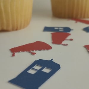 May include: Blue and red paper cutouts of a police box and a spaceship, scattered on a white surface. There are two cupcakes in the background.