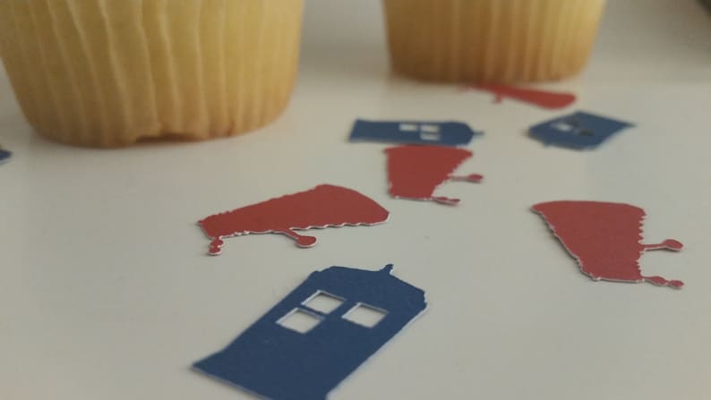 May include: Blue and red paper cutouts of a police box and a spaceship, scattered on a white surface. There are two cupcakes in the background.