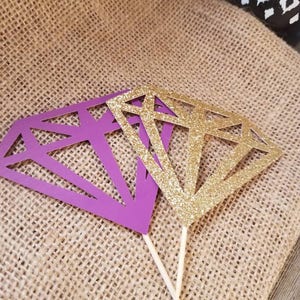 Pu&ograve; includere: Due topper per torta a forma di diamante, uno viola e uno dorato glitterato, su uno sfondo di juta.