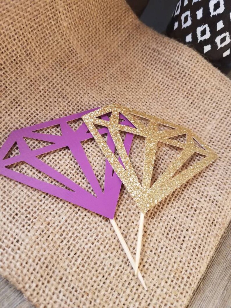 Pu&ograve; includere: Due topper per torta a forma di diamante, uno viola e uno dorato glitterato, su uno sfondo di juta.