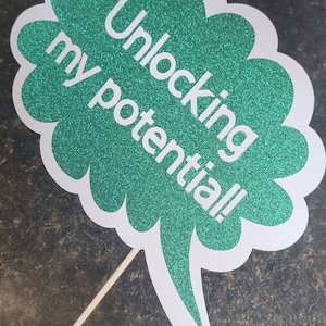 May include: A green glitter speech bubble with the text "Unlocking my potential!" printed in white. The speech bubble is attached to a wooden stick.
