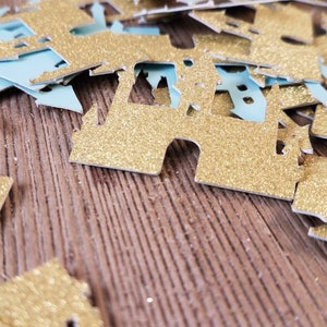 May include: Gold glitter castle confetti on a wood background. The confetti is in the shape of a castle with a pointed roof and towers.