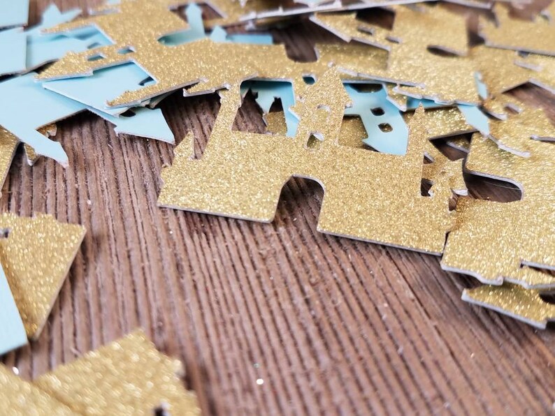 May include: Gold glitter castle confetti on a wood background. The confetti is in the shape of a castle with a pointed roof and towers.