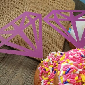 Pu&ograve; includere: Due ritagli di carta a forma di diamante viola su ciambelle con codette rosa, gialle e blu.