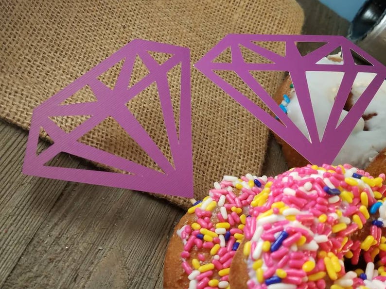 Pu&ograve; includere: Due ritagli di carta a forma di diamante viola su ciambelle con codette rosa, gialle e blu.