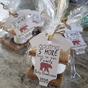 May include: A white tag with the text "adding S' MORE love to our Family Baby Gunderson 2020" and a red and black plaid bear graphic. The tag is attached to a wrapped s'more treat.