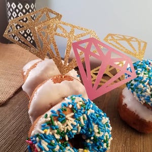 Pu&ograve; includere: Quattro ciambelle decorate con glassa bianca e codette colorate. Le ciambelle sono sormontate da ritagli di carta a forma di diamante con glitter dorati e rosa.
