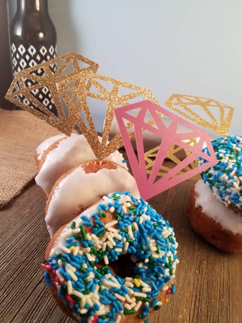 Pu&ograve; includere: Quattro ciambelle decorate con glassa bianca e codette colorate. Le ciambelle sono sormontate da ritagli di carta a forma di diamante con glitter dorati e rosa.