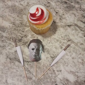 May include: Three cupcake toppers featuring a white and red swirl cupcake, a white mask with a black outline, and a silver knife with a brown handle.