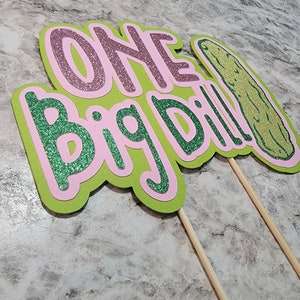 Op de afbeelding: Groene en roze glitterpapier-uitknipsels met de woorden "One Big Dill!" en een groene augurkvorm. De uitknipsels zijn bevestigd aan houten stokjes.