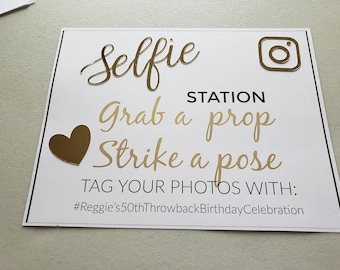 May include: A white sign with black text that reads "Selfie Station Grab a prop Strike a pose TAG YOUR PHOTOS WITH: #Reggie's50thThrowbackBirthdayCelebration". The sign has gold accents, including a heart, an Instagram logo, and the words "Selfie", "Grab", "Strike", and "pose".