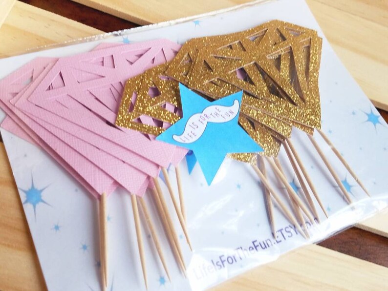 Pu&ograve; includere: Un pacchetto di topper per cupcake a forma di diamante con glitter rosa e oro, con un topper a forma di stella blu che dice "Life is for the fun" con un baffo.