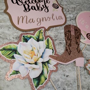 May include: Three pink and brown baby shower decorations. The first decoration is a sign that says "Welcome Baby Magnolia". The second decoration is a pink and brown boot. The third decoration is a white magnolia flower with green leaves.
