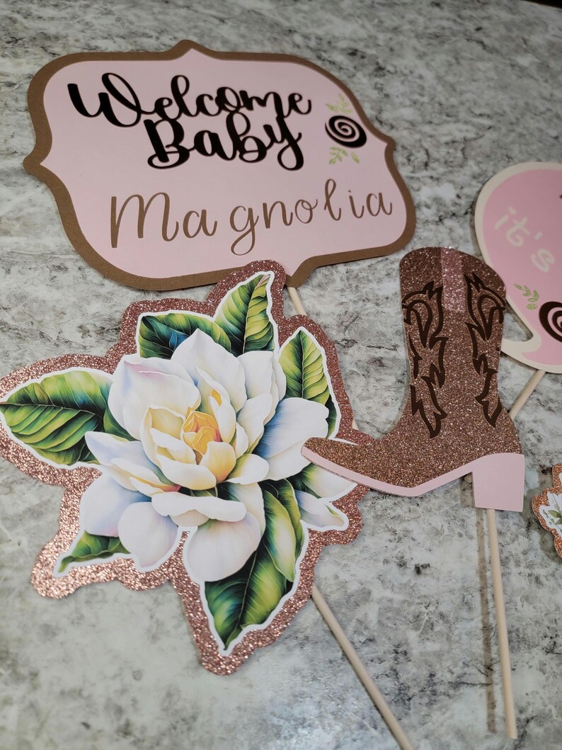May include: Three pink and brown baby shower decorations. The first decoration is a sign that says "Welcome Baby Magnolia". The second decoration is a pink and brown boot. The third decoration is a white magnolia flower with green leaves.