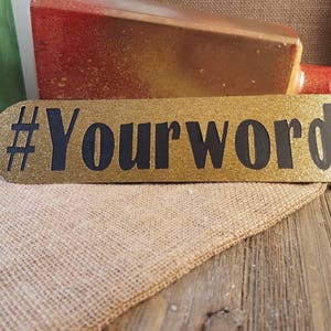 May include: A gold glitter sign with the text "#Yourwords" in black. The sign is oval-shaped and rests on a burlap cloth and a wooden surface. Other decorative items are visible in the background.