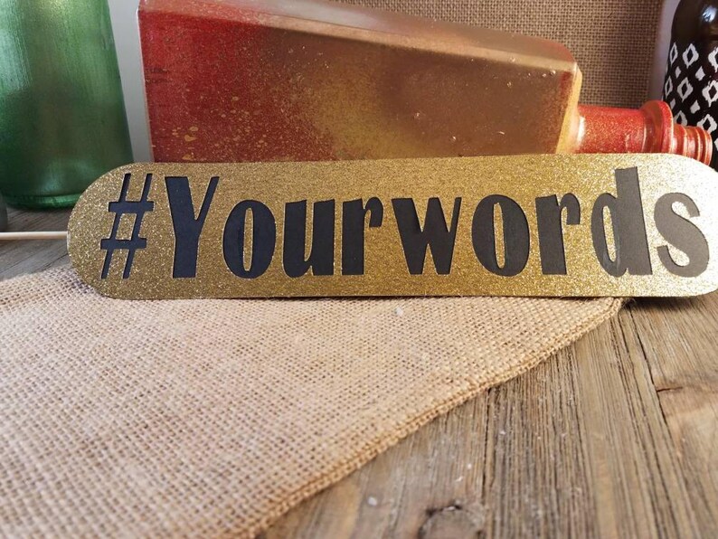 May include: A gold glitter sign with the text "#Yourwords" in black. The sign is oval-shaped and rests on a burlap cloth and a wooden surface. Other decorative items are visible in the background.