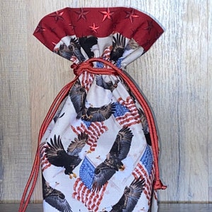 May include: A white drawstring bag with a red and white American flag pattern featuring bald eagles. The bag has a red drawstring and a red fabric top with white stars.