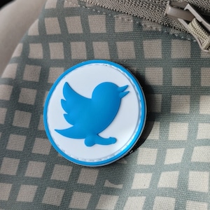 May include: A blue and white round patch with the Twitter bird logo. The patch is attached to a brown and white checkered fabric.