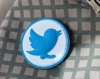 May include: A blue and white round patch featuring the Twitter bird logo. The patch is attached to a brown and white checked fabric.