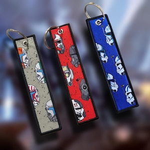 May include: Three rectangular keychains with metal rings. One is gray with colorful helmet designs, another is red with black and white helmet designs, and the third is blue with white helmet designs. Each keychain has a black border.