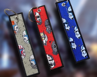 May include: Three rectangular keyrings with metal hoops. One is grey with colourful helmet designs, another is red with black and white helmet designs, and the third is blue with white helmet designs. Each keyring has a black border.