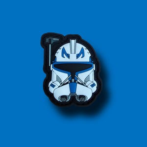 Puede incluir: Un parche de casco blanco y azul con borde negro, con un diseño detallado. El casco tiene una visera y marcas distintivas, sobre un fondo azul. El parche es probablemente de goma o material similar.