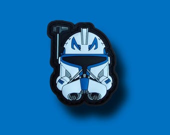 May include: A white and blue helmet patch with a black border, featuring a detailed design. The helmet has a distinctive visor and markings, set against a blue background. The patch is likely made of rubber or a similar material.
