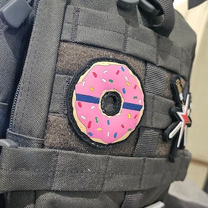 May include: Close-up of a tactical backpack with a pink donut patch. The donut has pink frosting, colorful sprinkles, and a blue line. Additional patches include a star and a badge. The backpack is dark gray.
