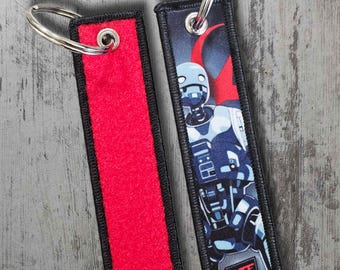 May include: Two rectangular keyrings. One is red with a black border and a silver key ring. The other keyring has a black border and features a blue and white robot graphic with the text "WSF". Both keyrings are on a grey surface.