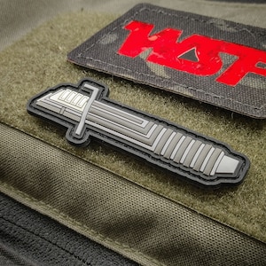May include: A black and gray tactical patch with a stylized image of a lightsaber. The patch is designed to be attached to a tactical vest or bag.