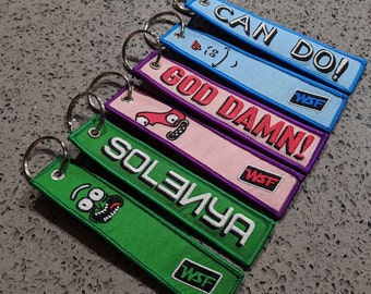 May include: Six colourful keychains with different phrases and designs. The keychains are made of fabric and have a metal ring. The phrases on the keychains are "Can Do!", "God Damn!", "Solemya", and "(oo)".