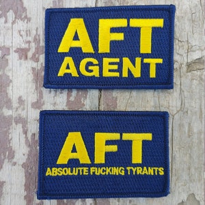 May include: Two blue embroidered patches with yellow lettering. The top patch reads "AFT AGENT". The bottom patch reads "AFT ABSOLUTE FUCKING TYRANTS".