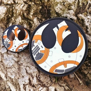 Könnte beinhalten: Zwei runde, schwarz-weiße Emaille-Pins mit orangefarbenem Design. Der größere Pin zeigt das Symbol der Rebellenallianz mit einem BB-8-Design. Der kleinere Pin ist eine kleinere Version des BB-8-Designs.
