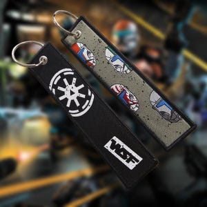 Pode incluir: Dois chaveiros bordados. Um é preto com um símbolo branco e as letras "WSF". O outro é cinza com imagens repetidas de capacetes em vermelho, azul e verde. Ambos têm argolas prateadas.