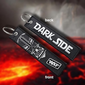 May include: Two black embroidered keychains with silver key rings. One keychain reads "DARK SIDE" in white lettering. The other keychain has a white graphic and the letters "WSF". The background is a blurred image of red and orange.