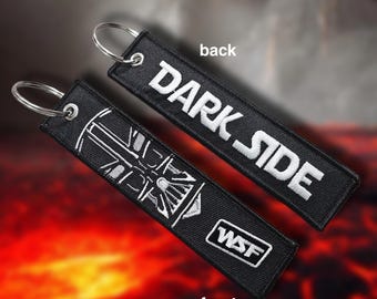 May include: Two black embroidered keychains with silver key rings. One keychain reads "DARK SIDE" in white lettering. The other keychain has a white graphic and the letters "WSF". The background is a blurred image of red and orange.