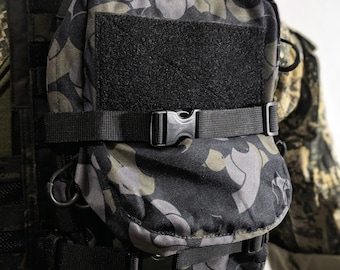 May include: A camouflage-patterned tactical vest accessory with a black hook-and-loop patch, adjustable straps, and a secure buckle. The pouch is designed to attach to a tactical vest.