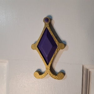 May include: A gold-colored door hook with a purple diamond-shaped design in the center. The hook is mounted on a white door.