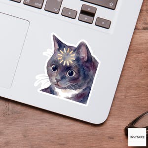 Può includere: Un adesivo ad acquerello di un gatto nero con un disegno a forma di sole dorato sulla fronte. Il gatto ha grandi occhi espressivi e segni bianchi sul petto. L'adesivo è posto su un laptop bianco.