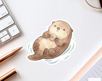 May include: A watercolour sticker of a cute otter floating in blue water, holding a seashell. The otter is brown and tan with a sweet expression. The sticker is on a white surface with a keyboard, pen, glasses, and notebook.