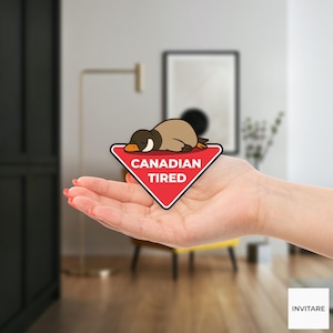 Pu&ograve; includere: Un adesivo triangolare rosso con le parole "CANADIAN TIRED" e un'anatra dei cartoni animati che riposa sopra. L'anatra &egrave; marrone e beige con un becco arancione. L'adesivo &egrave; tenuto in una mano.