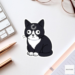Puede incluir: Una pegatina de gato en blanco y negro con un símbolo de carga sobre los ojos. El gato tiene ojos grandes y redondos y una expresión de sorpresa. La pegatina está sobre una superficie blanca, con un teclado, un bolígrafo y un cuaderno visibles.