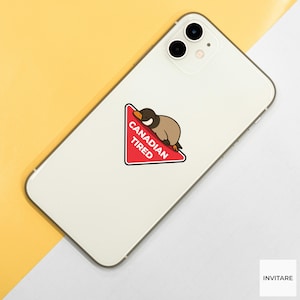 Pu&ograve; includere: Uno smartphone bianco con un adesivo raffigurante un'anatra dei cartoni animati che riposa su un triangolo rosso. Il triangolo ha le parole "CANADIAN TIRED" in bianco. Il telefono &egrave; su una superficie gialla e bianca.