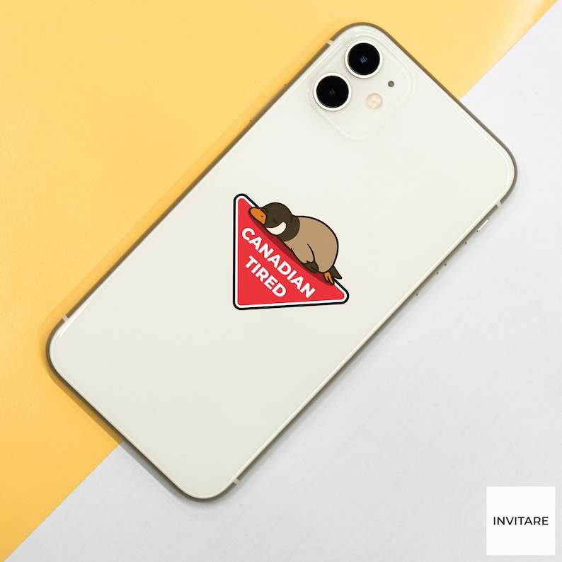 Pu&ograve; includere: Uno smartphone bianco con un adesivo raffigurante un'anatra dei cartoni animati che riposa su un triangolo rosso. Il triangolo ha le parole "CANADIAN TIRED" in bianco. Il telefono &egrave; su una superficie gialla e bianca.