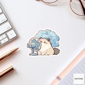 May include: A die-cut sticker featuring a fluffy cream-colored cat with dark brown ears and tail sitting next to a small light blue electric desk fan. Behind the cat is a large, soft, light blue cloud shape. The illustration has a white border.