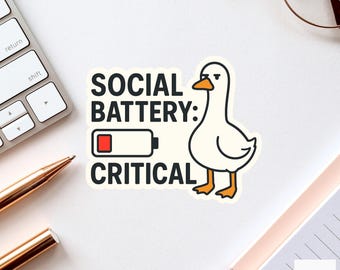 Pode incluir: Um adesivo branco com o texto "SOCIAL BATTERY: CRITICAL" e uma ilustração de pato de desenho animado. O pato tem bico e pés amarelos. Um ícone de bateria fraca está ao lado do texto. O adesivo está em uma superfície branca.