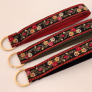 May include: Three keyrings with floral embroidered straps. The straps are black with red and gold floral patterns. Each keyring has a gold-coloured ring.